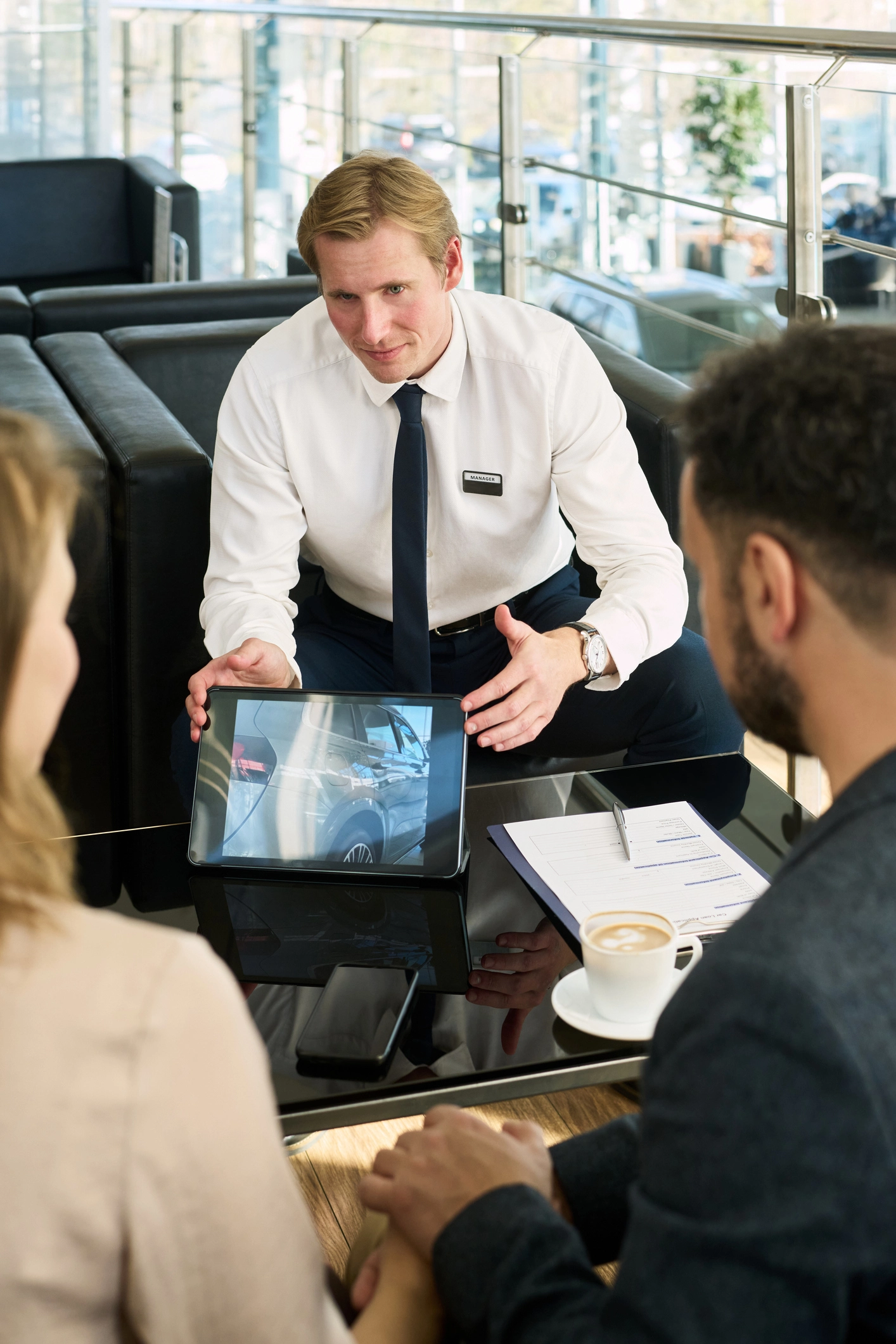 Verkaufsgespräch Mitarbeiter präsentiert zwei Kunden im Beratungsgespräch in einem Autohaus digitale Produktinformationen auf einem Tablet.